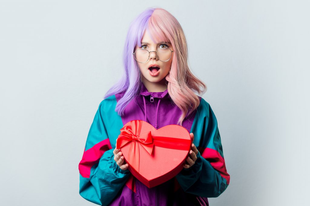 Beautiful yandere girl cosplayer with purple hair and 80s tracksuit hold heart shape gift box on gray background