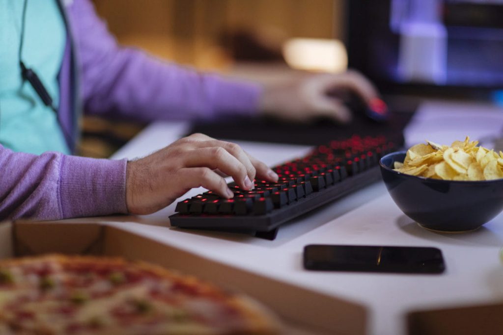Close up shot of male hands typing on keyboard with snacks and other food within easy reach